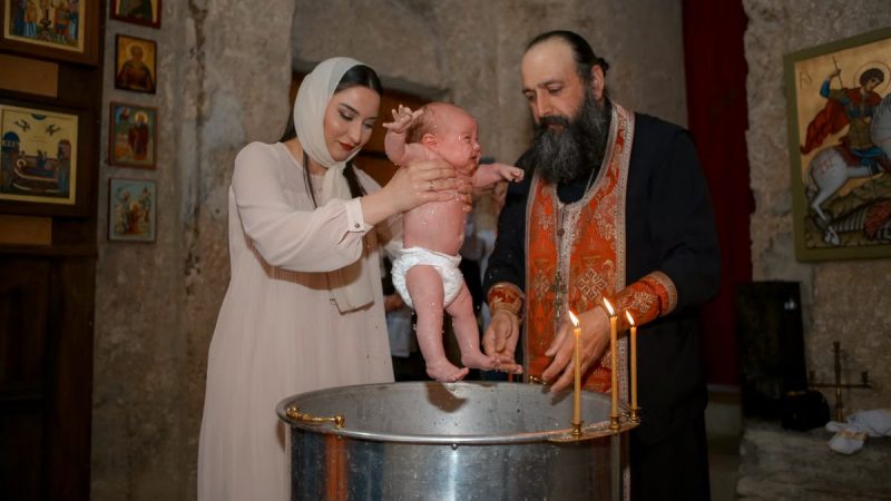 A baby being baptized by a priest and woman in a traditional church setting.